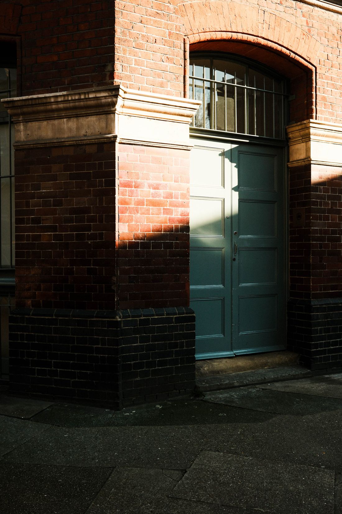 A quiet brick building corner with a arched doorway and dark green door, lit by soft afternoon sunlight, empty of people.