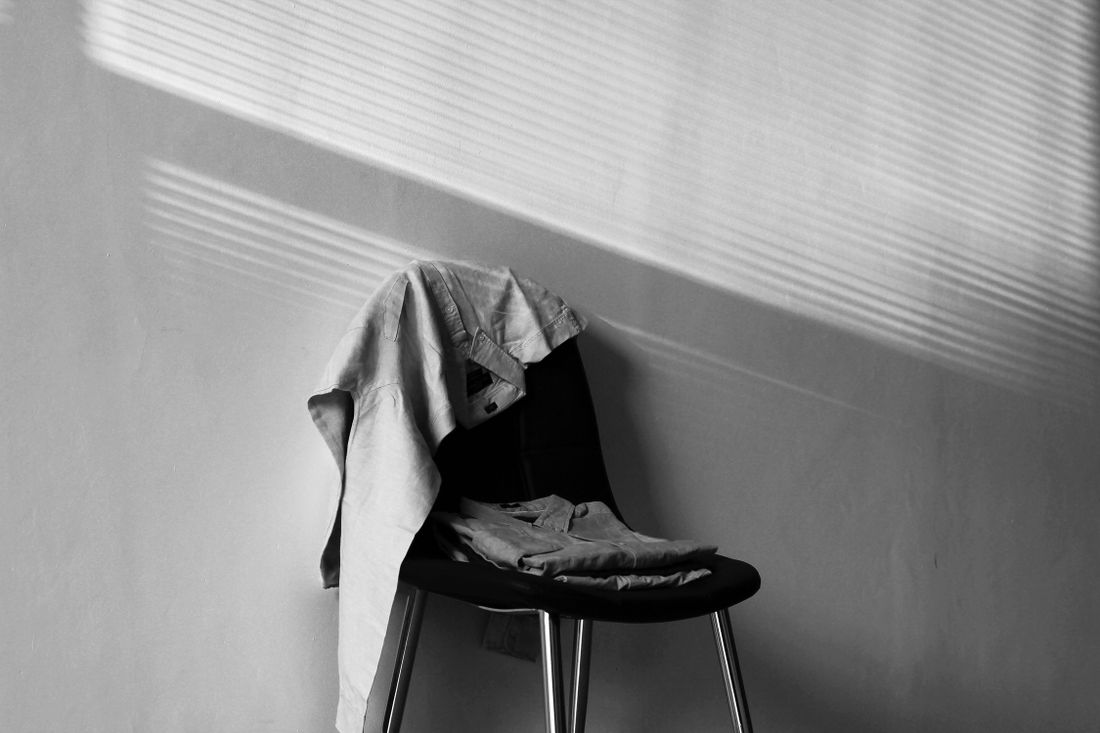 A black and white photo of a shirt draped over a chair back and folded clothing resting on the seat, with striped window-light shadows on the wall behind.