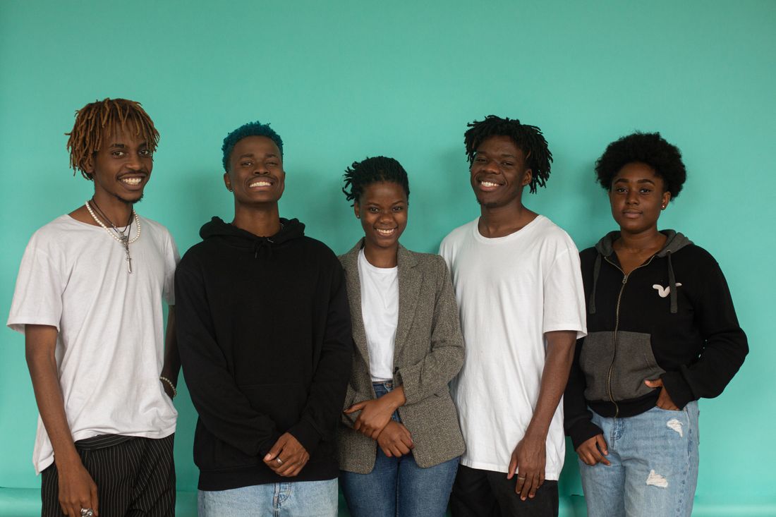 Five people of varying heights standing together against a solid teal backdrop, arranged in a loose offset cluster rather than a straight line