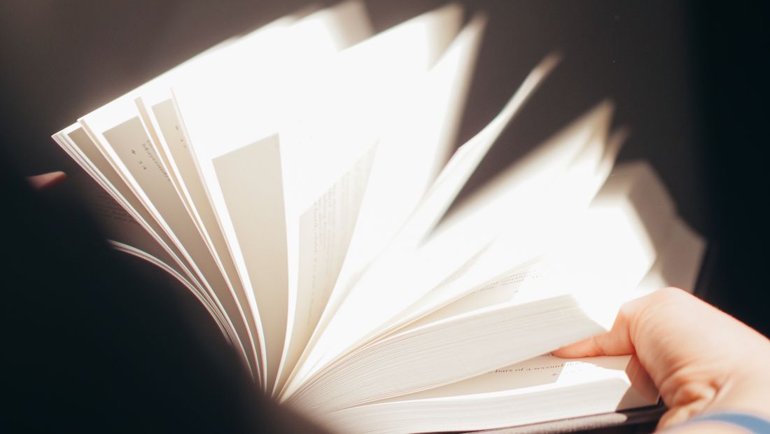 A hand fans open the pages of a book, backlit so the paper glows and the printed text is illegible.