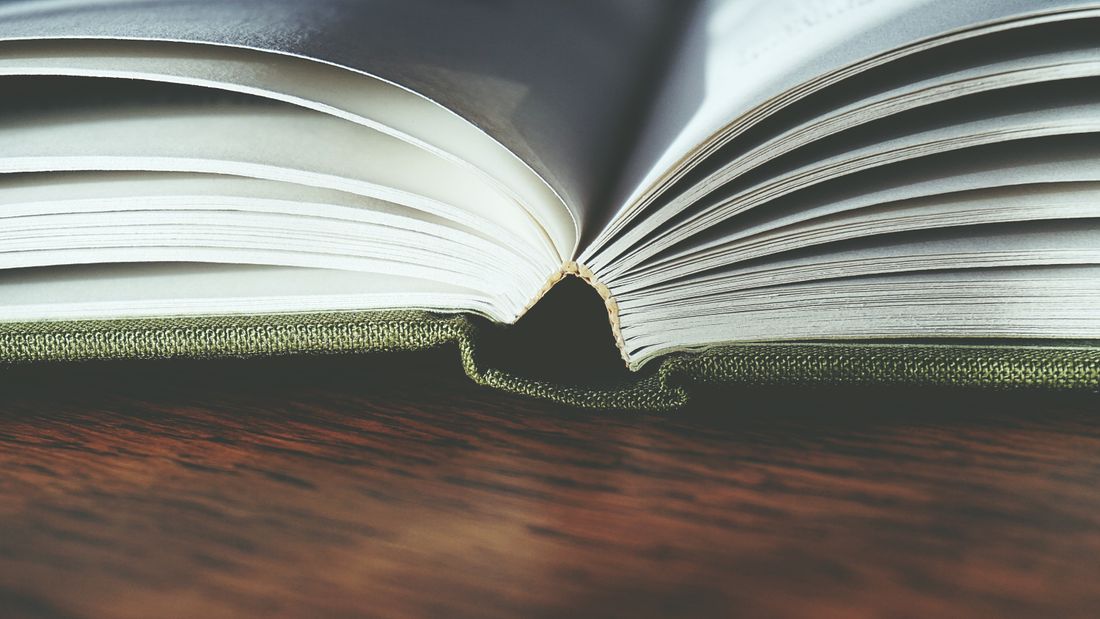 Close low-angle view of an open hardcover book on a wooden table, showing the curved gutter and cloth binding where the pages meet the spine.