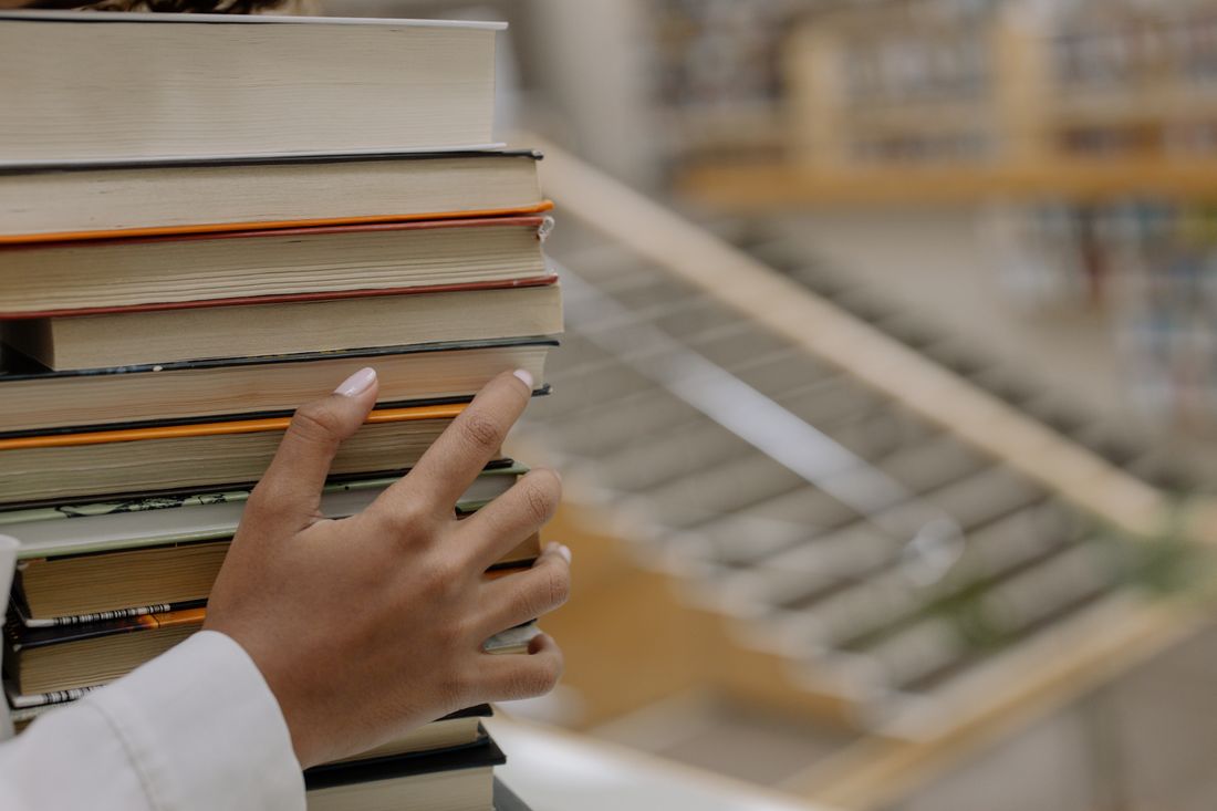 A hand holding the side of a stack of hardcover books, foredges and spines visible, blurred shelves in the background.
