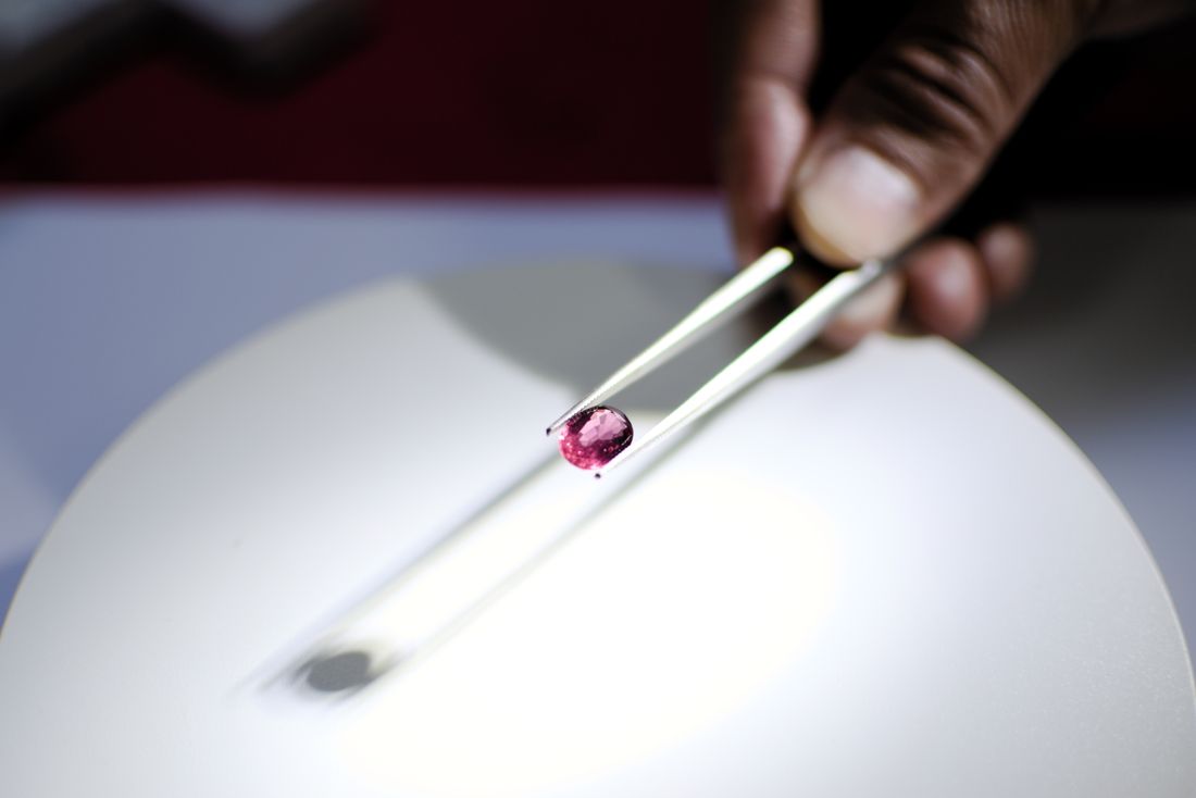 A hand using tweezers to hold a small red gemstone above a lit white surface