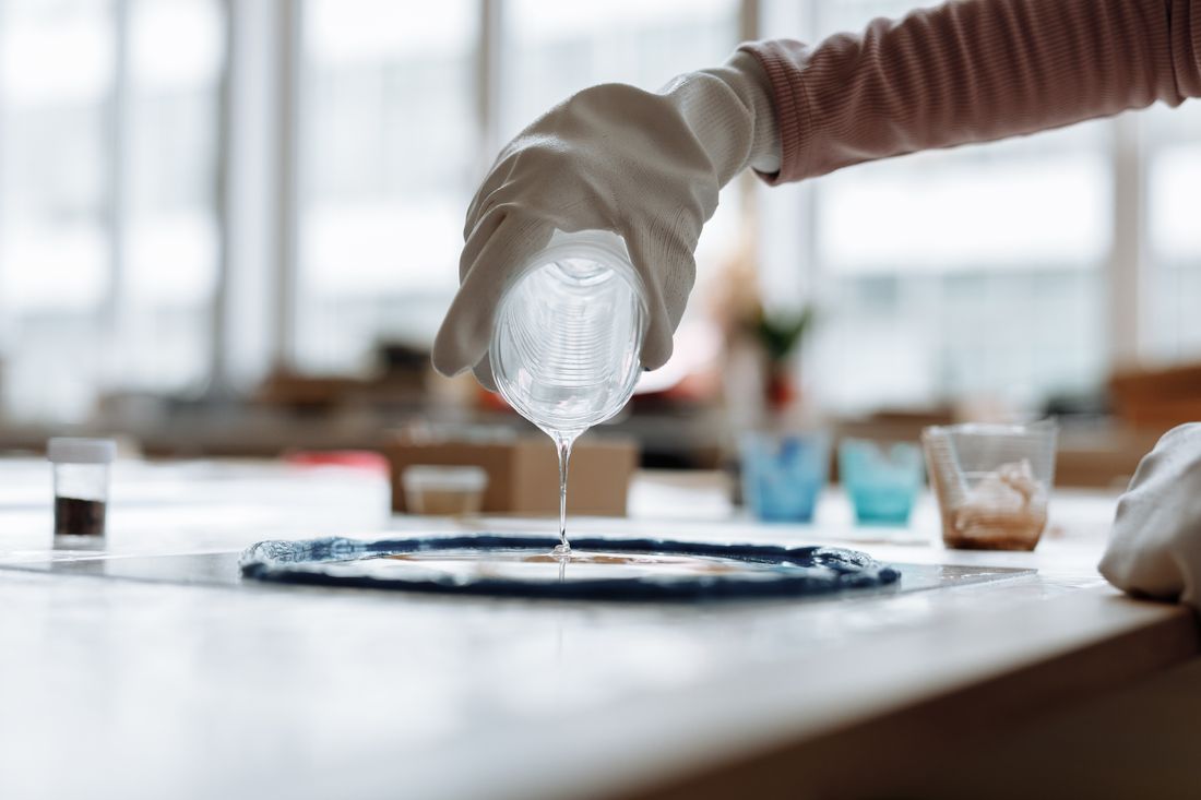 A gloved hand pouring clear resin onto a mold on a worktable, showing the material's glossy, reflective surface