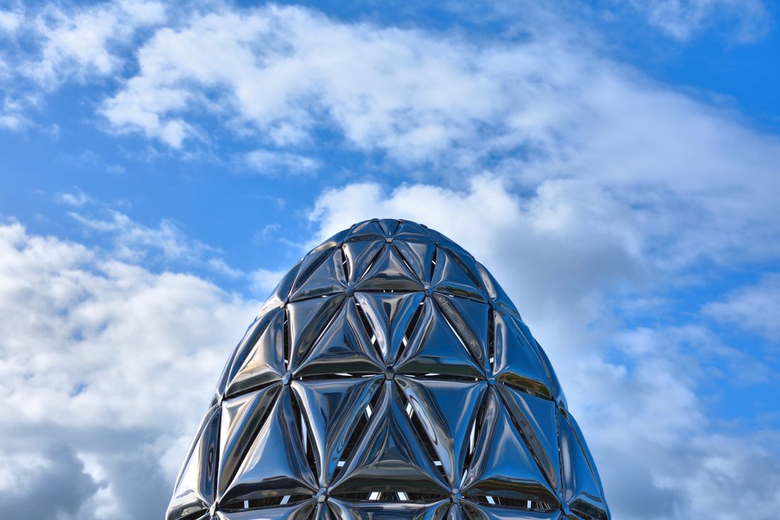 A polished mirror-finish sculpture reflecting a cloudy blue sky, showing how a reflective surface picks up everything around it
