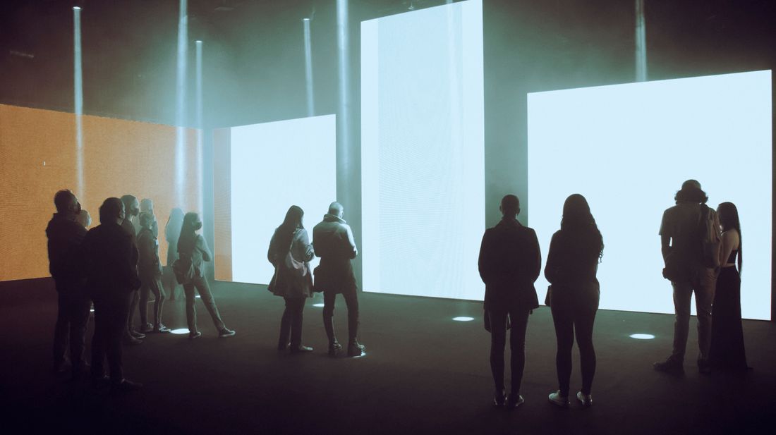 A darkened exhibition room where several large projected screens glow pale blue, with visitors standing in front of them as silhouettes.