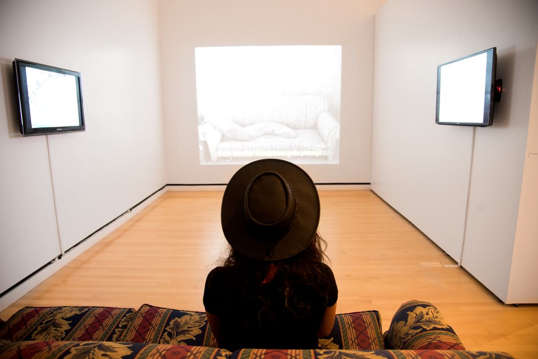A small gallery room with a flat screen mounted on each side wall and a projection filling the far wall, seen past a seated viewer photographed from behind.