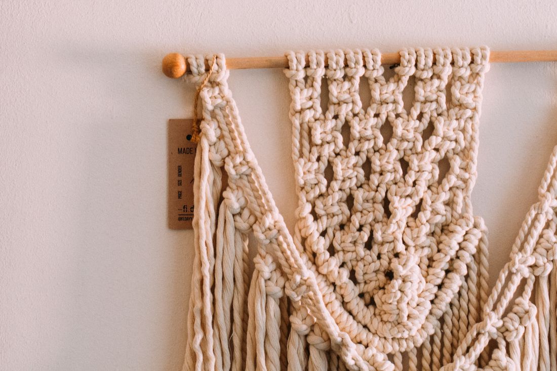 A handmade beige macrame wall hanging showing intricate knot structure and fringe against a plain wall
