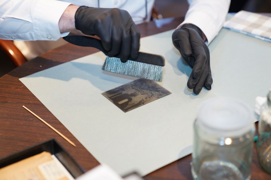 Gloved hands using a soft brush to carefully handle a small paper photograph during preservation work