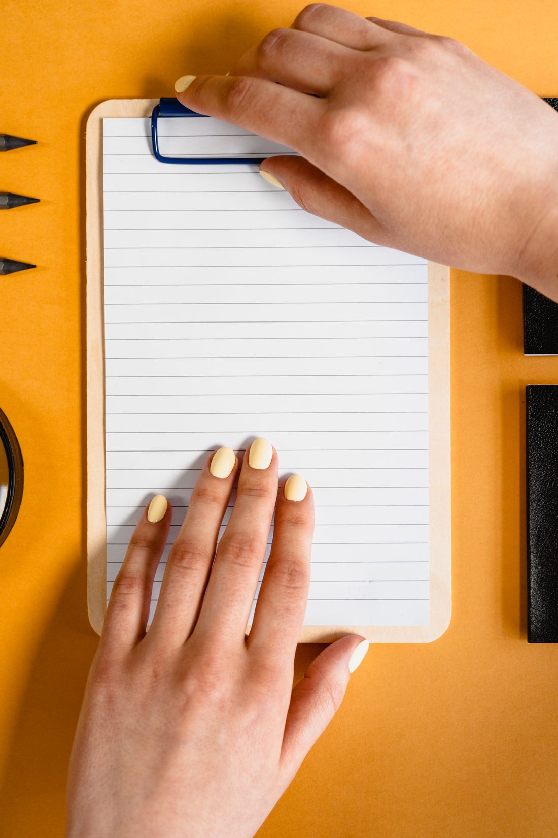 Overhead view of two hands resting on a blank lined notepad on a clipboard at a desk.