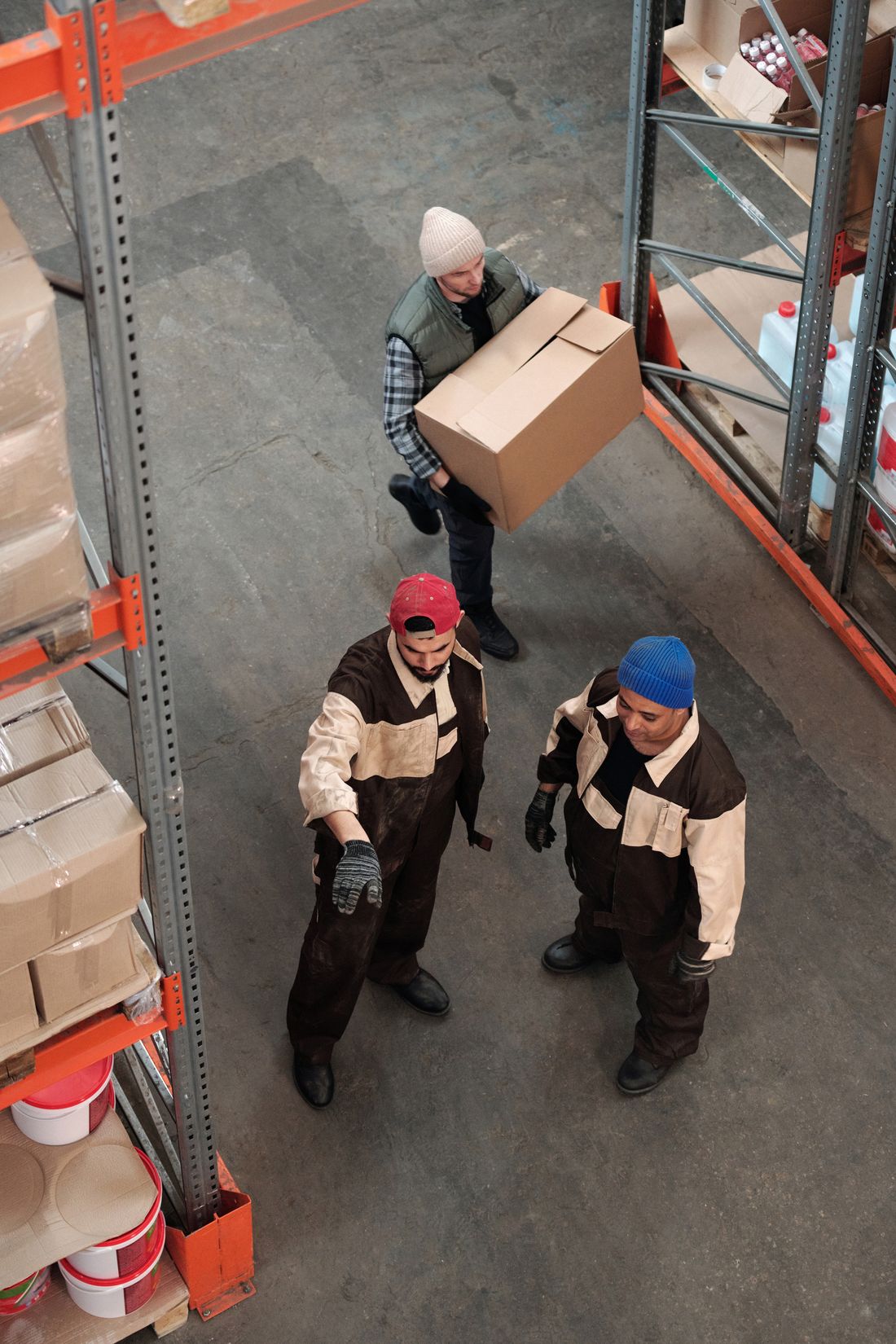 Warehouse staff carrying a cardboard box past two coworkers on an indoor floor lined with storage racks.
