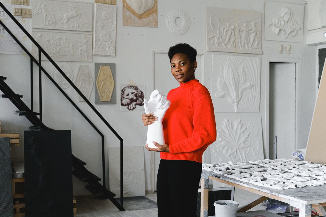 A person in a red sweater standing at a worktable in a plaster-walled studio holding a small object