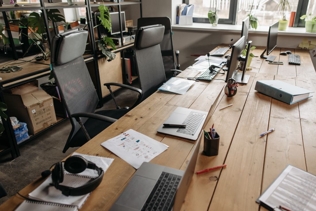A shared office desk with papers, a laptop and headphones beside a window