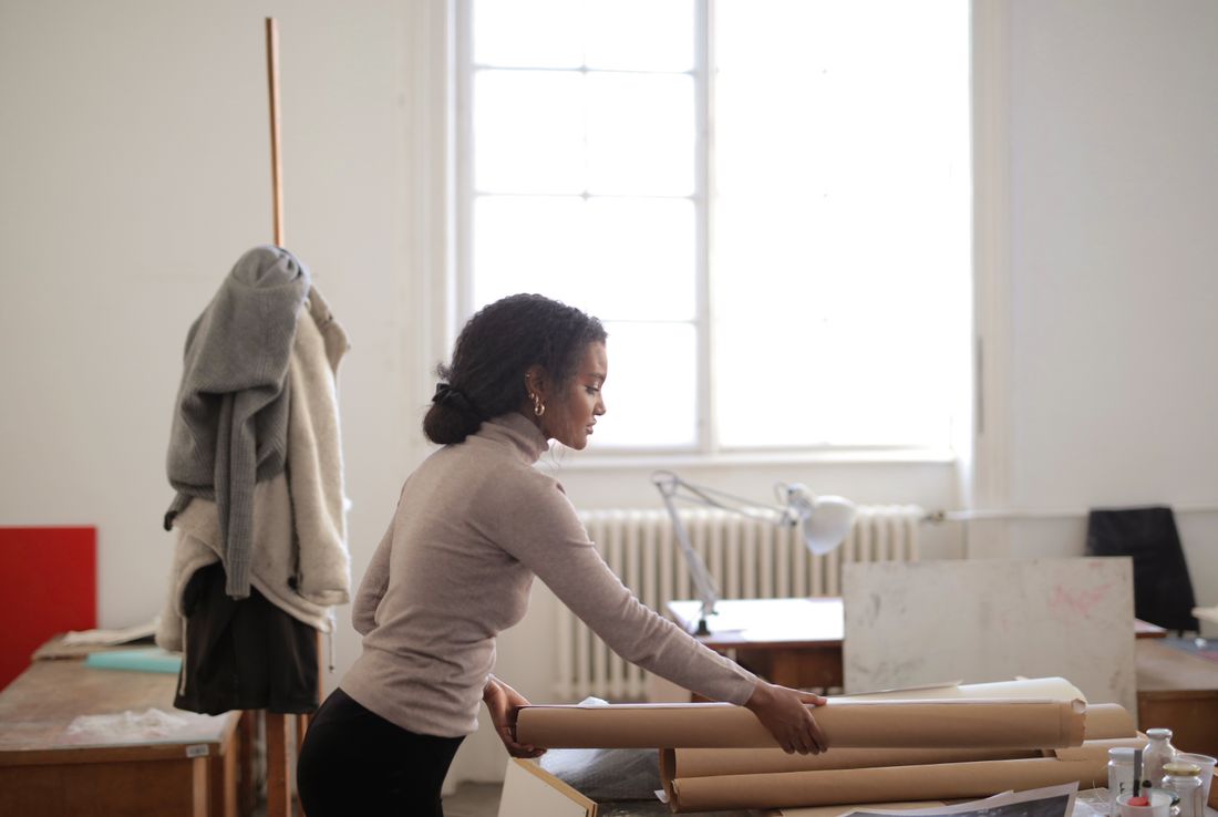 A woman seen from the side unrolls a length of kraft paper on a worktable in a sunlit studio, surrounded by rolls of paper and a hanging sweater.