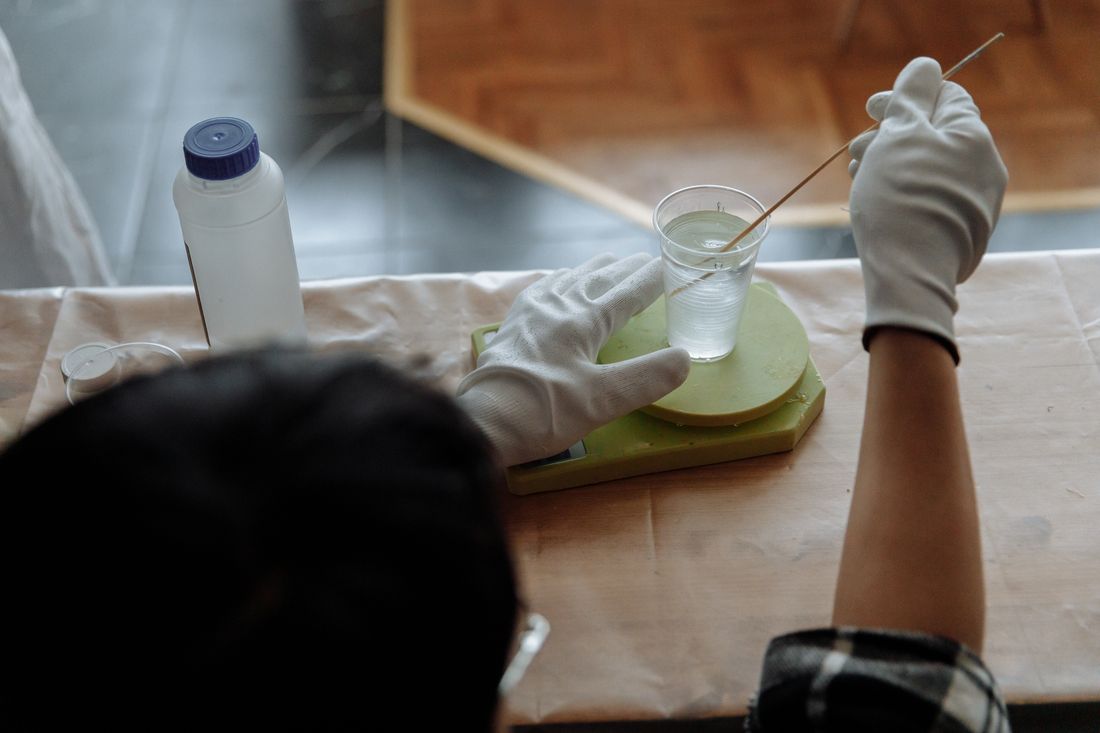 Close-up of hands in white work gloves stirring a clear liquid with a thin tool over a glass, a second person's head visible but out of focus in the foreground.