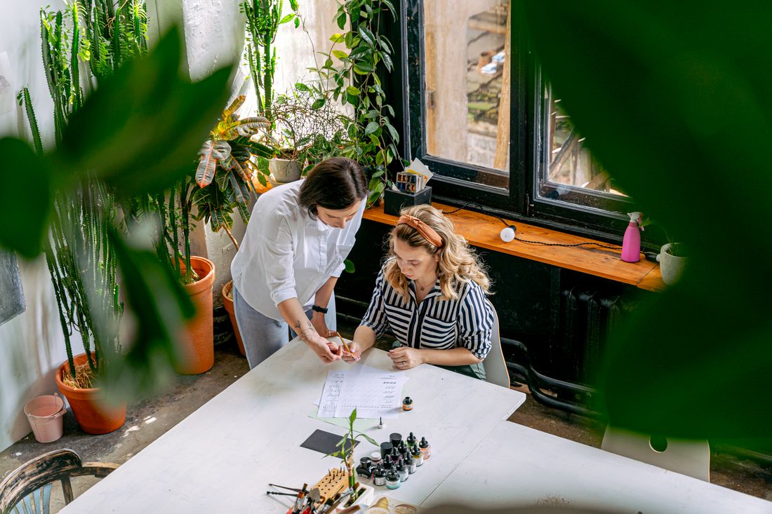 Two people lean over a table together in a plant-filled studio, both looking down at a sheet of paper and small glass vials between them.