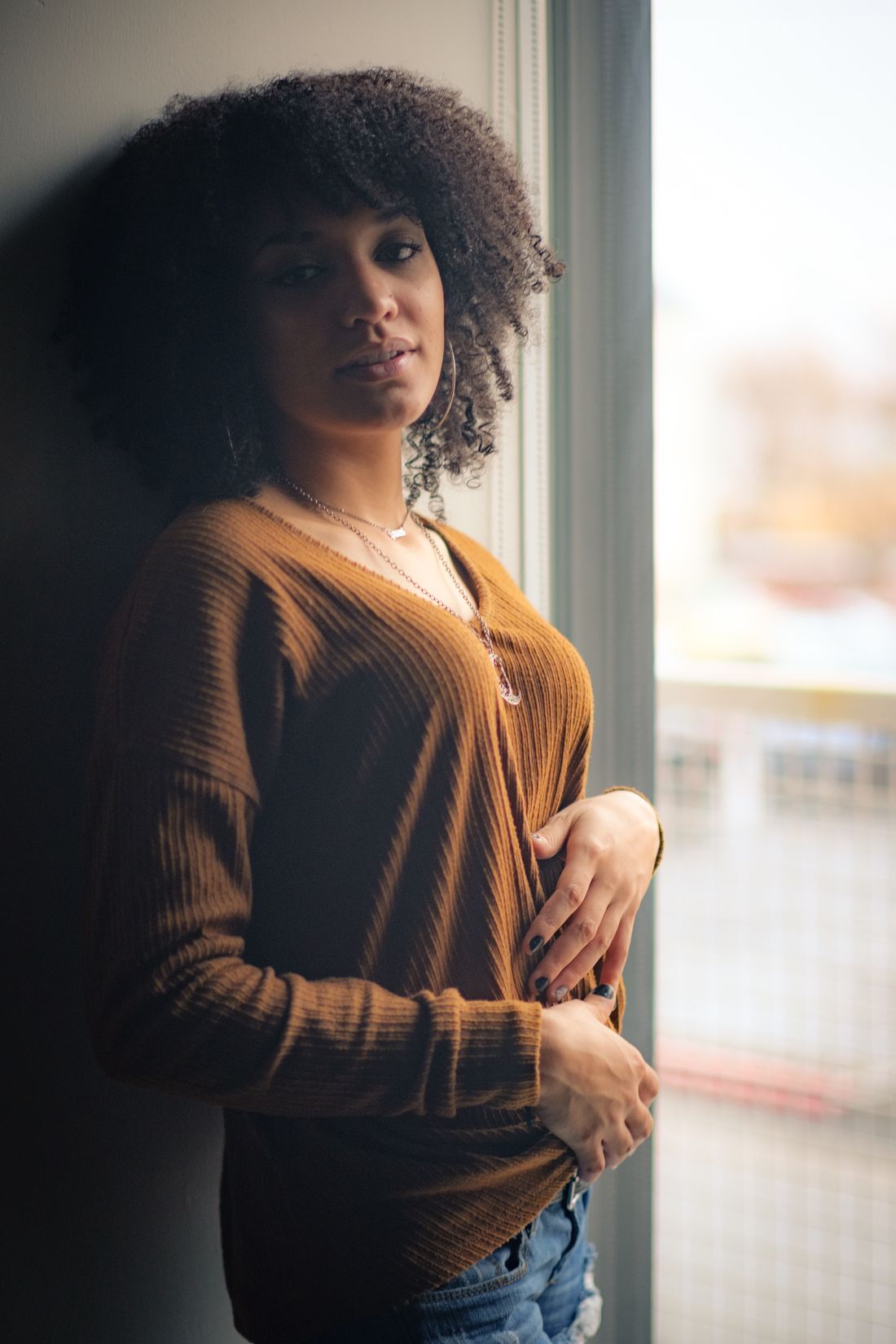 A woman in a warm-toned sweater leans against a wall by a window, looking back over her shoulder with a thoughtful expression.