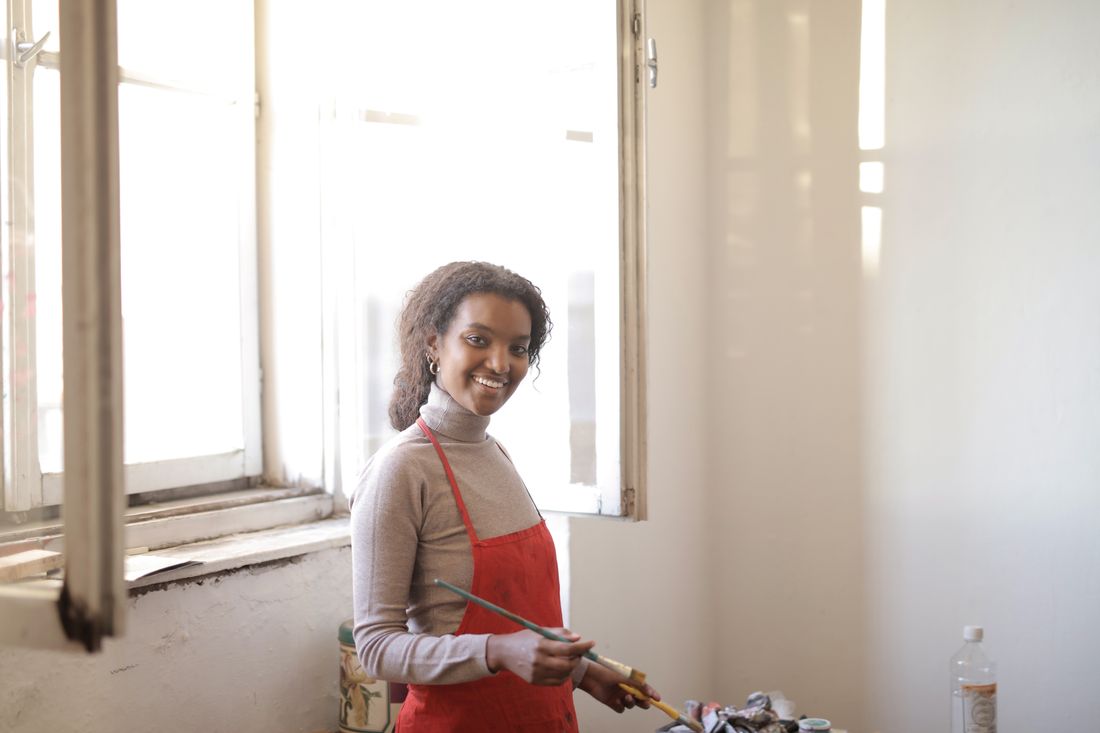 A woman in a red apron stands smiling by a bright studio window, holding a paintbrush over a palette of paints.