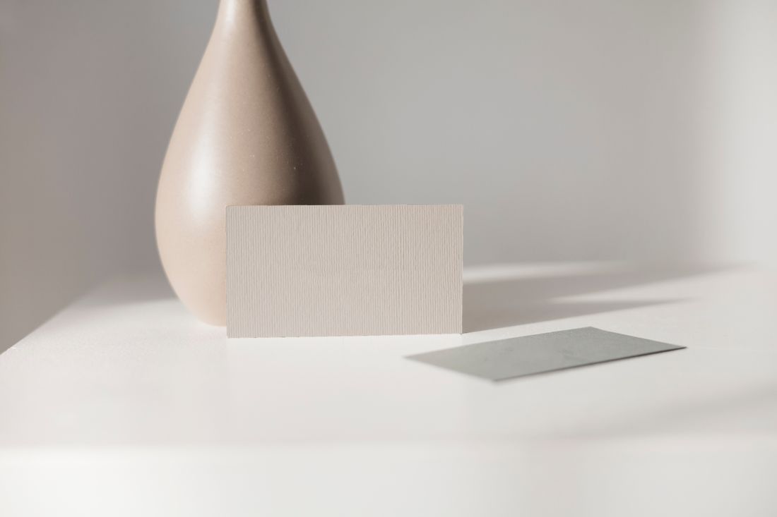 A blank card propped upright against a ceramic vase on a pale tabletop, with a second card lying flat nearby.