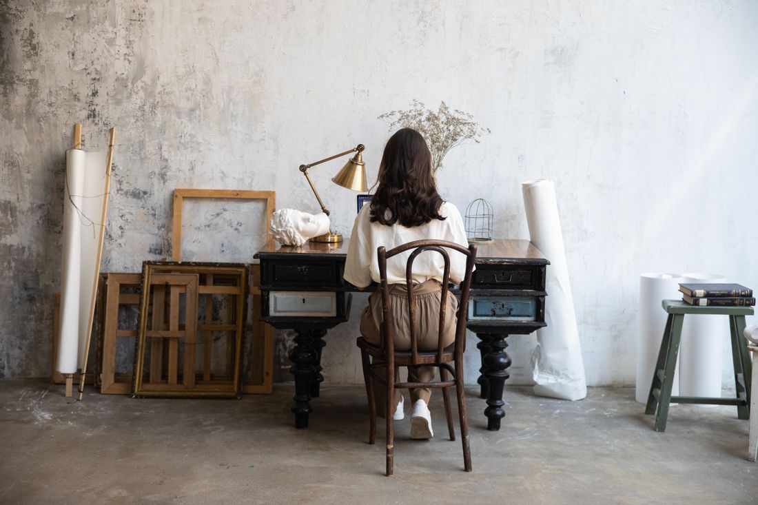 A woman seen from behind, seated at a vintage desk against a plain studio wall, surrounded by empty picture frames and rolled paper, working with hands near a small desk lamp.