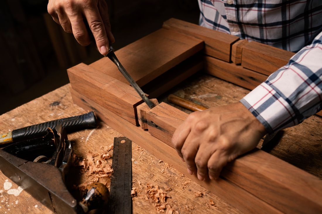 Close crop of two hands gripping a wood chisel over a workbench, mid-motion, wood shavings scattered around, no face or head in frame.