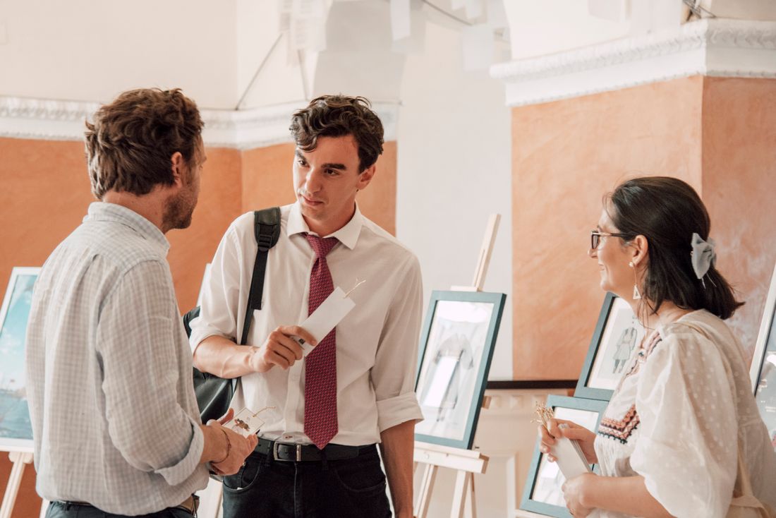 Three people in conversation in a gallery, framed artwork on easels around them