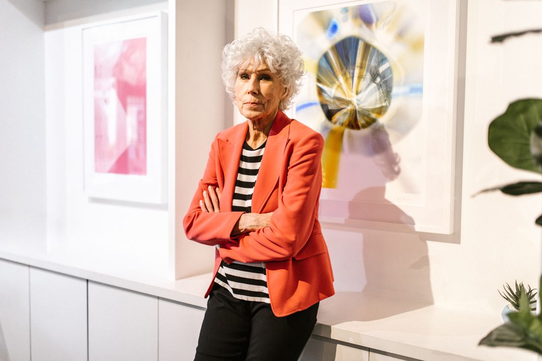 A woman in a red blazer stands with arms crossed in a gallery, paintings visible on the walls behind her