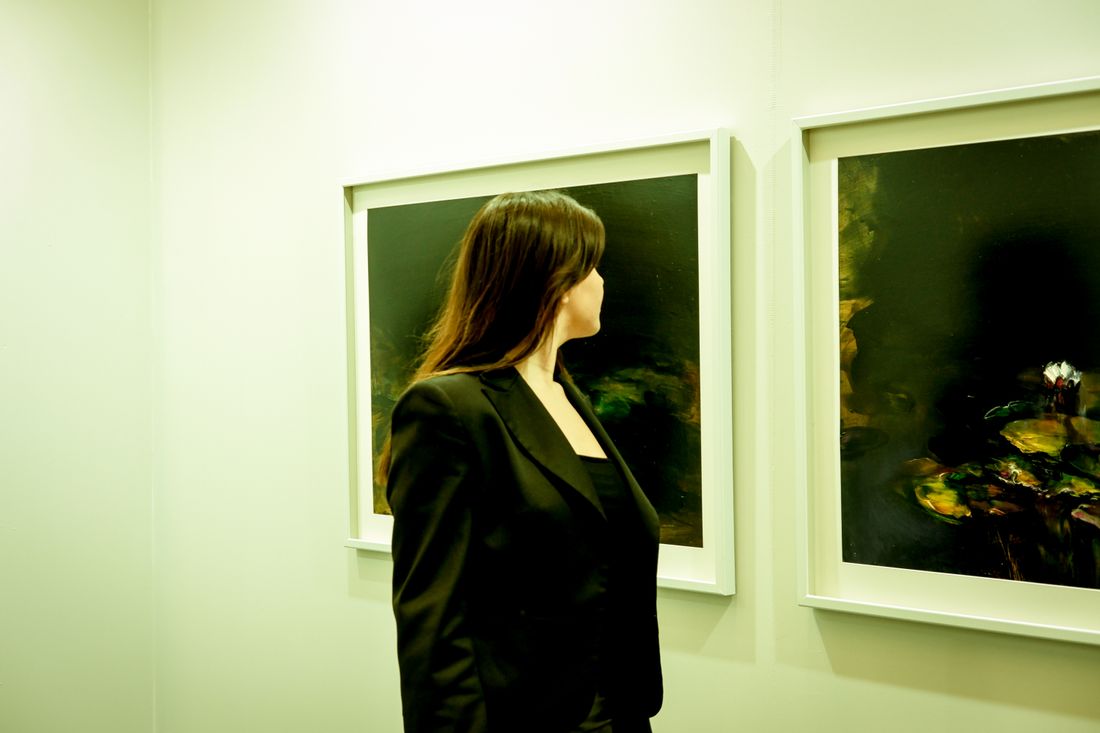 A woman in a dark blazer viewed in profile, looking at framed paintings on a gallery wall