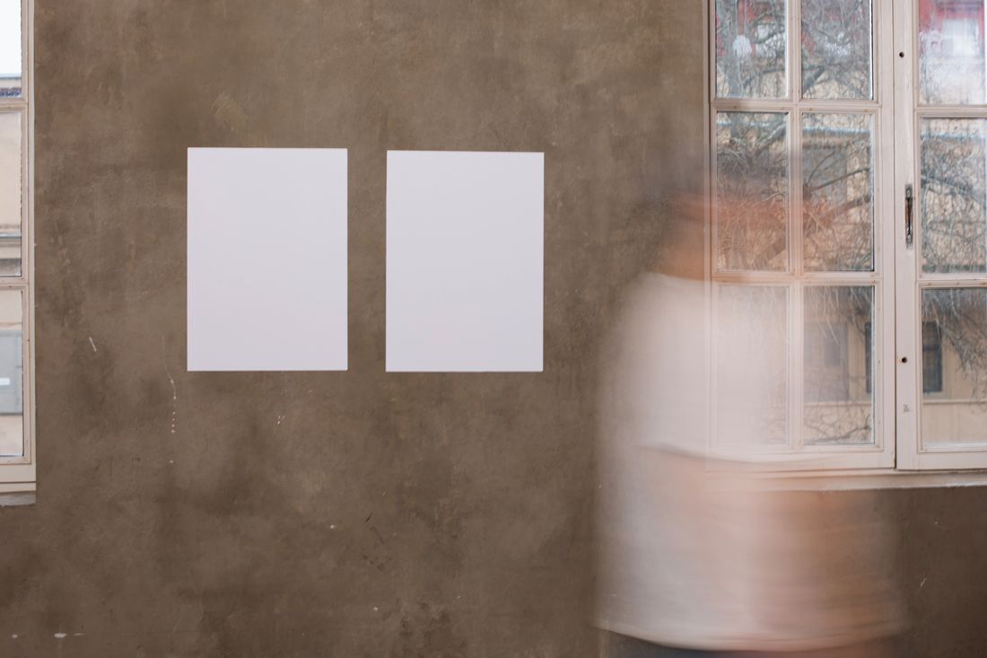 A blurred figure walks past two blank mockup frames on a textured wall near a window, caught mid-motion.