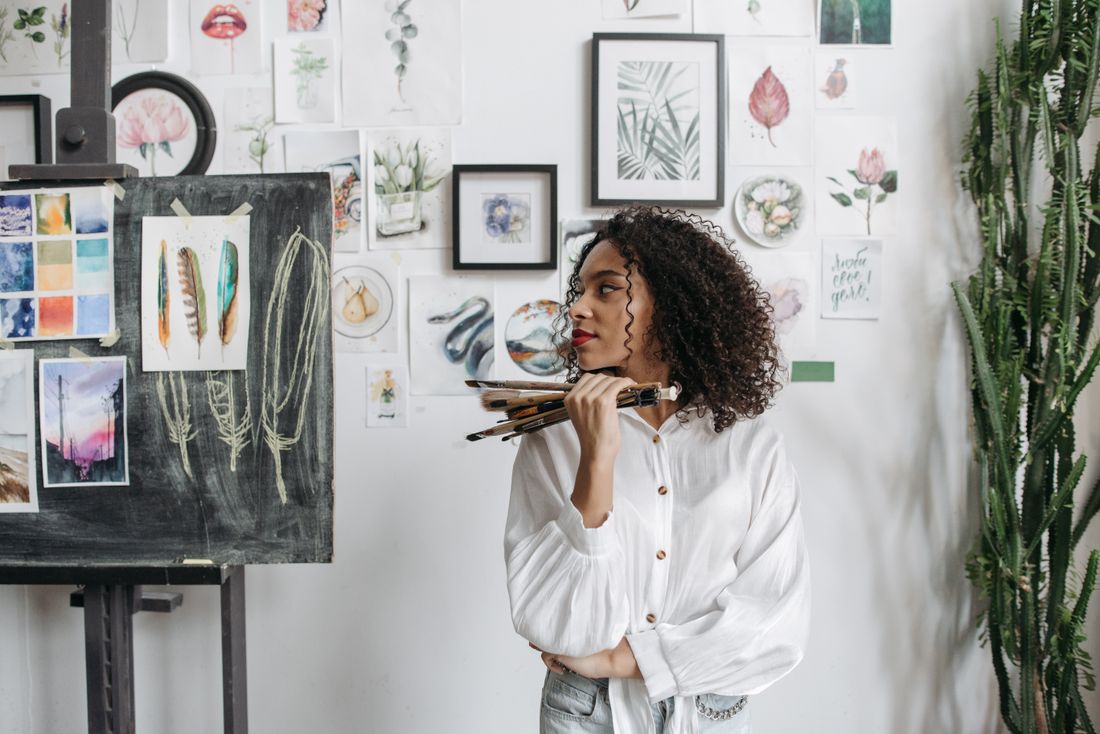 A painter holds a handful of brushes and looks away from the camera in a studio with reference images pinned to the wall.