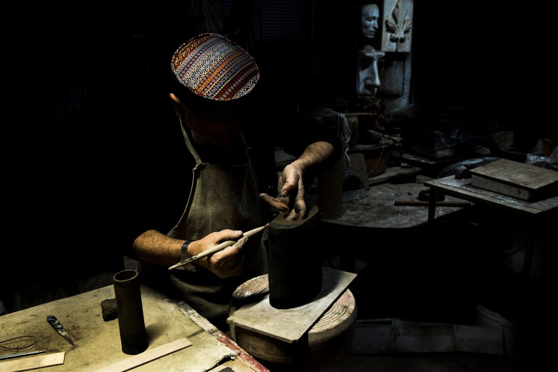 A craftsman works on a small object at a cluttered worktable, face partly visible under low workshop light.