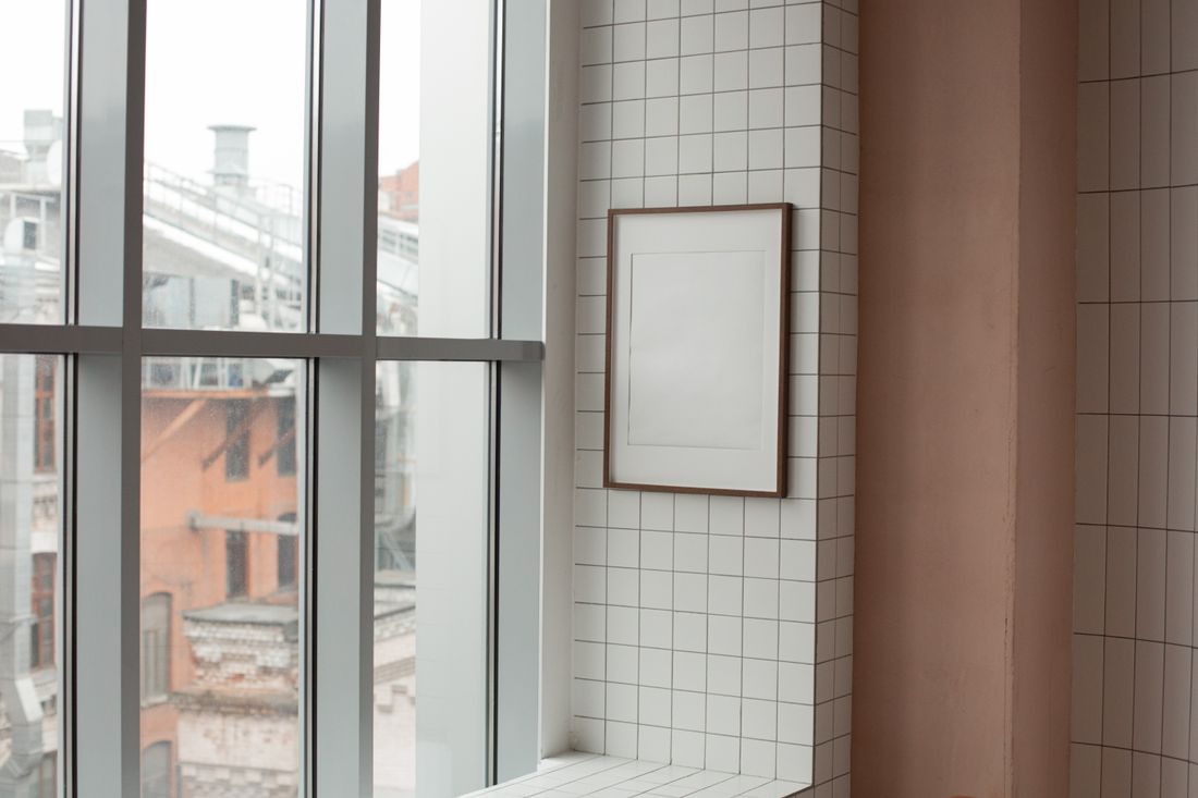 An empty picture frame on a tiled wall beside a large window, photographed from an angle rather than straight on.