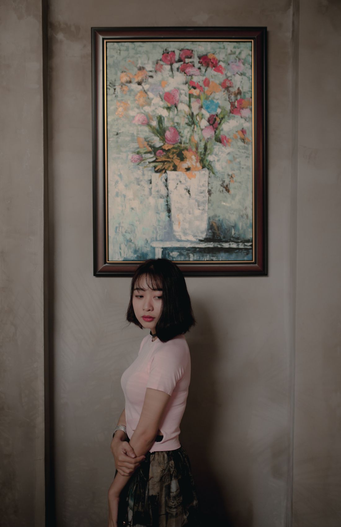 A woman stands beside a single framed floral still-life painting hanging on a plain wall