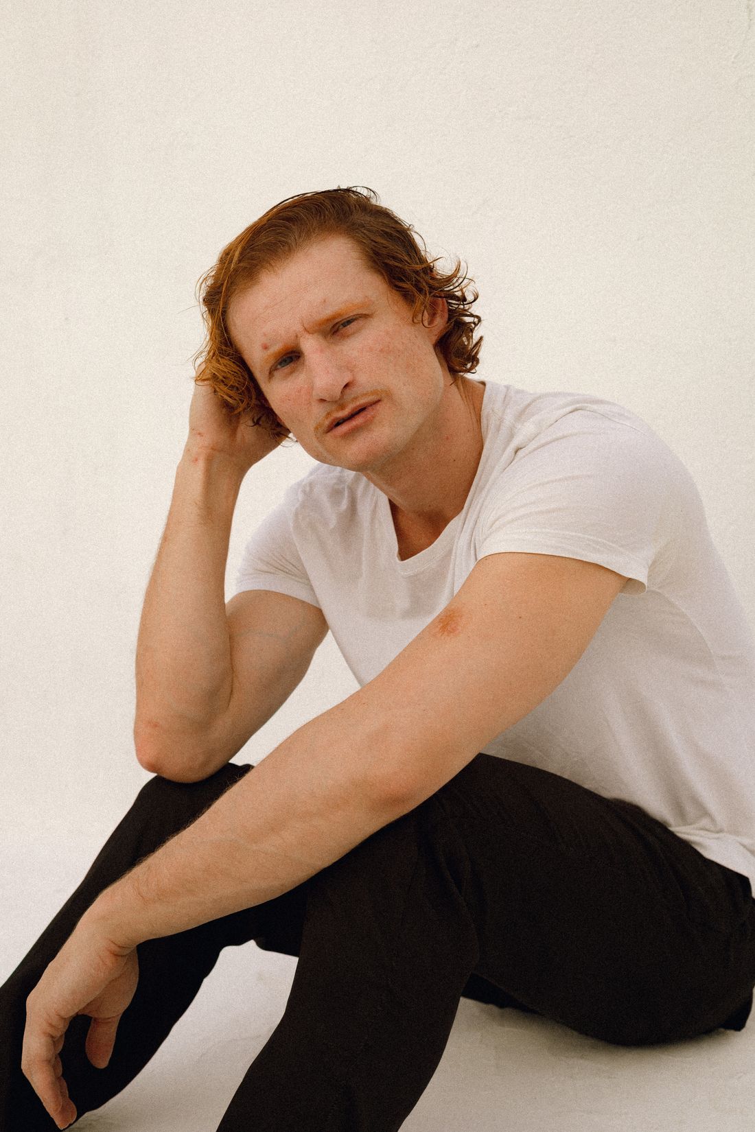 A portrait of a person sitting against a plain wall, one hand resting against their hair, looking off to the side with a thoughtful expression