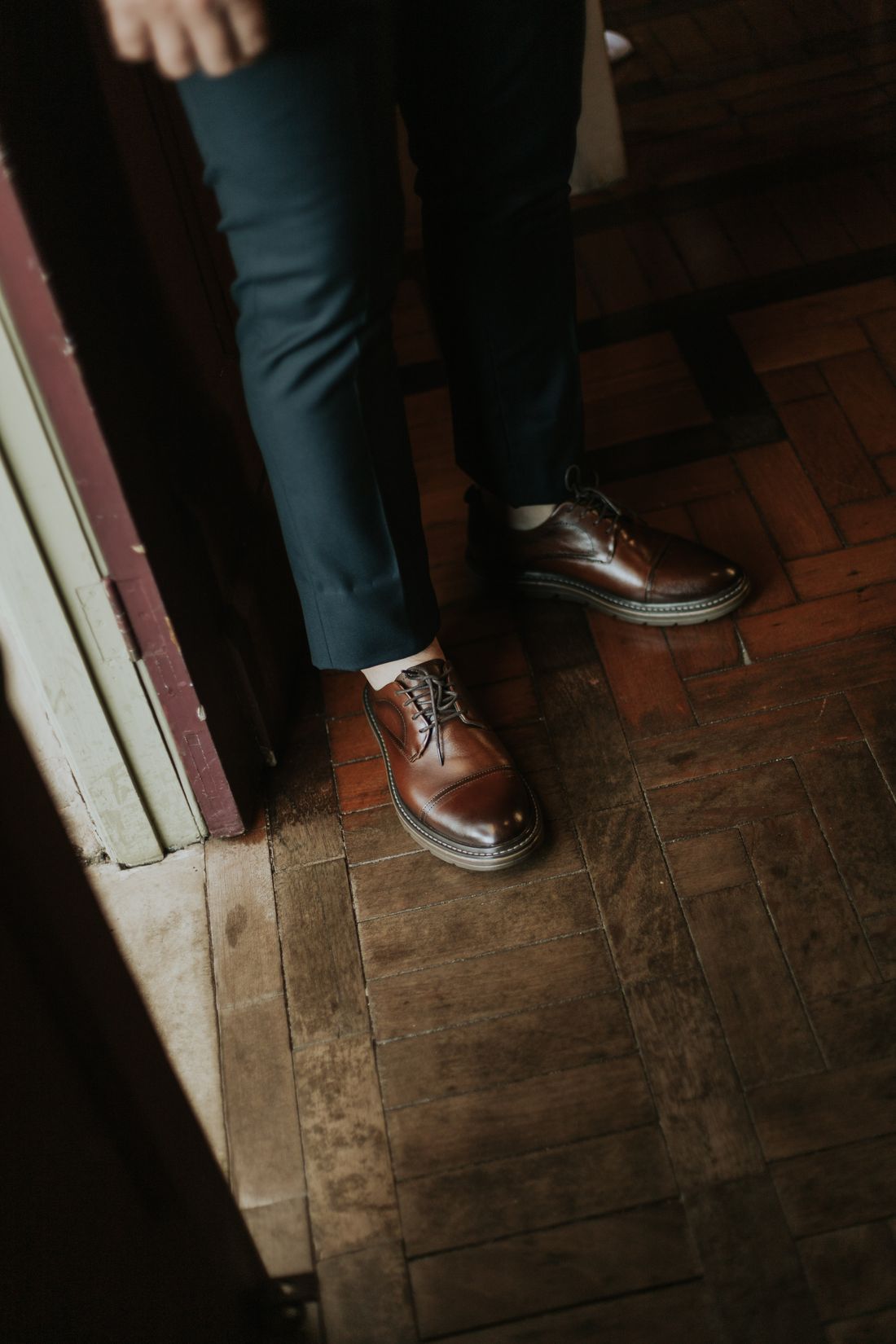 Close crop of a person's legs and brown leather dress shoes stepping across a wood floor near a doorway