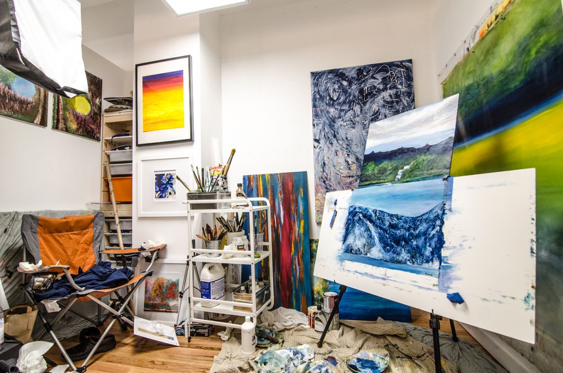 A working artist's studio with finished and in-progress paintings on shelves and an easel, paint supplies on a cart