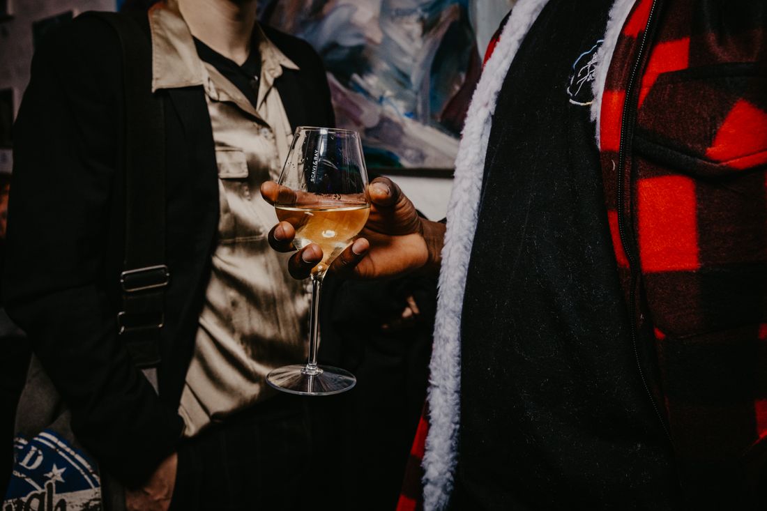 A hand holding a wine glass in front of a painting at a crowded gallery event, the artwork partly obscured