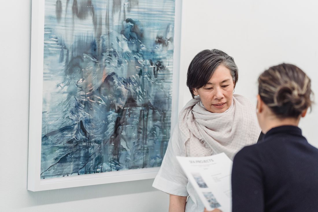 Two women in conversation beside an abstract painting, one holding printed materials