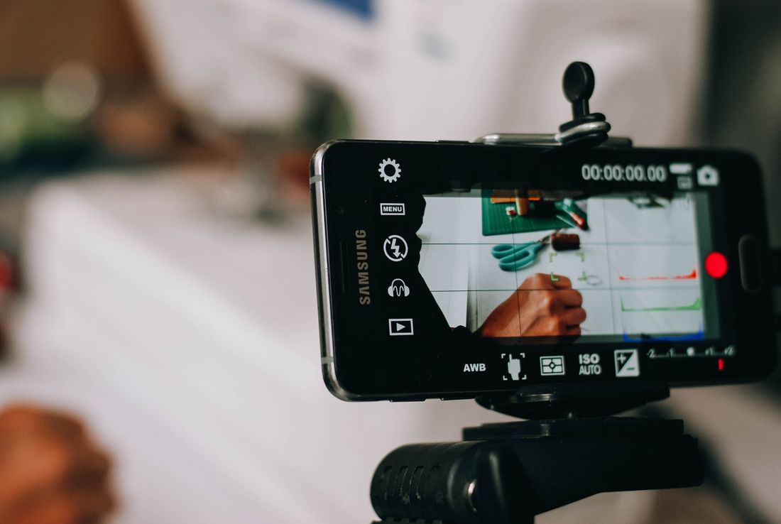 A phone or small camera mounted on a tripod, screen showing a close recording of hands working with materials on a table