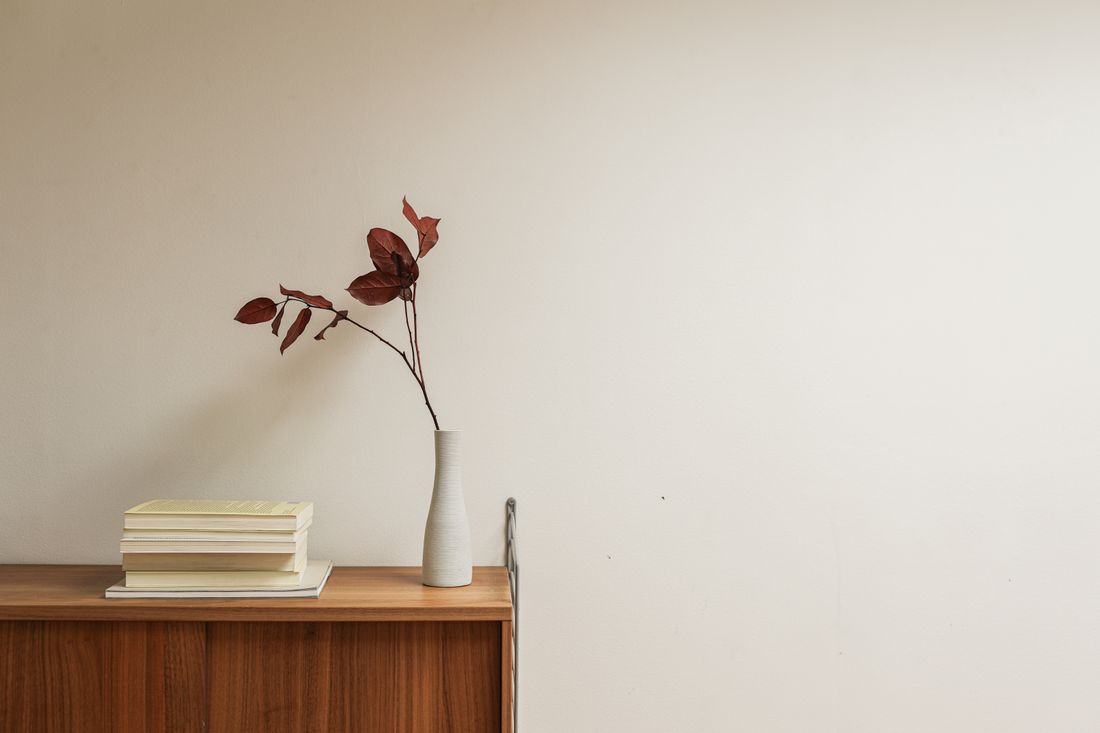 A ceramic vase holding a dried branch beside a small stack of books on a wooden sideboard against a plain wall.