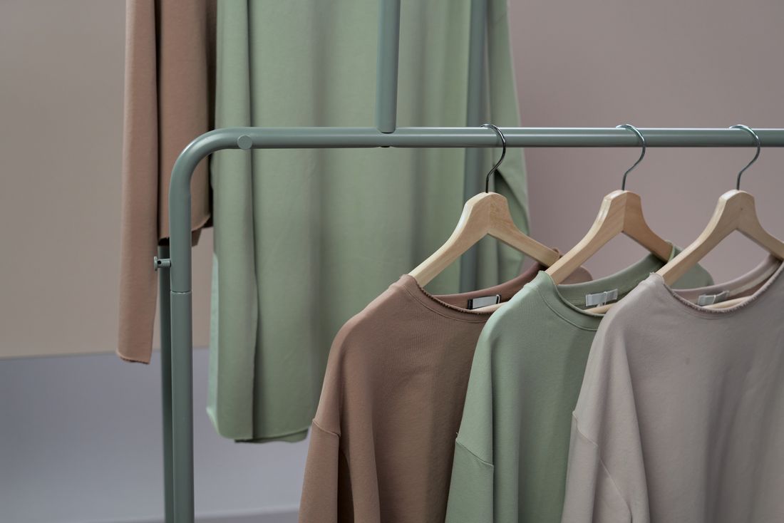 Three neutral-toned sweaters on wooden hangers on a clothing rack, with folded garments draped alongside.