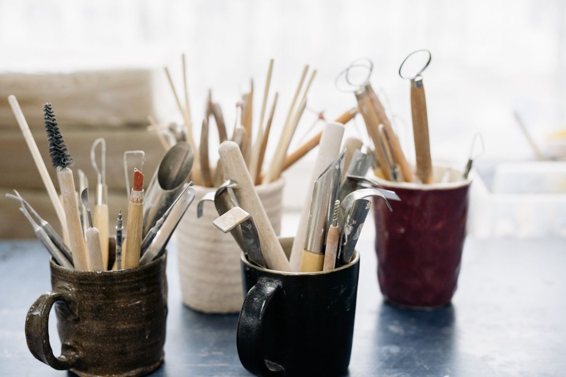 A row of well-used clay sculpting tools standing in ceramic mugs on a worktable, soft window light behind.