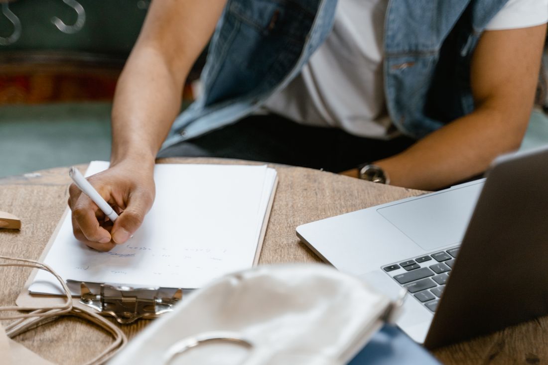 A seated person in a denim vest writing on paper clipped to a binder at a wooden table, the edge of an open laptop beside them