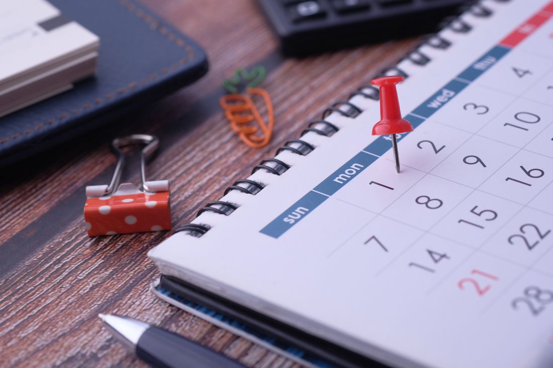 A spiral desk calendar on a wooden table with a red push pin marking a Monday date, a pen, a calculator, and a binder clip nearby, nobody in frame.