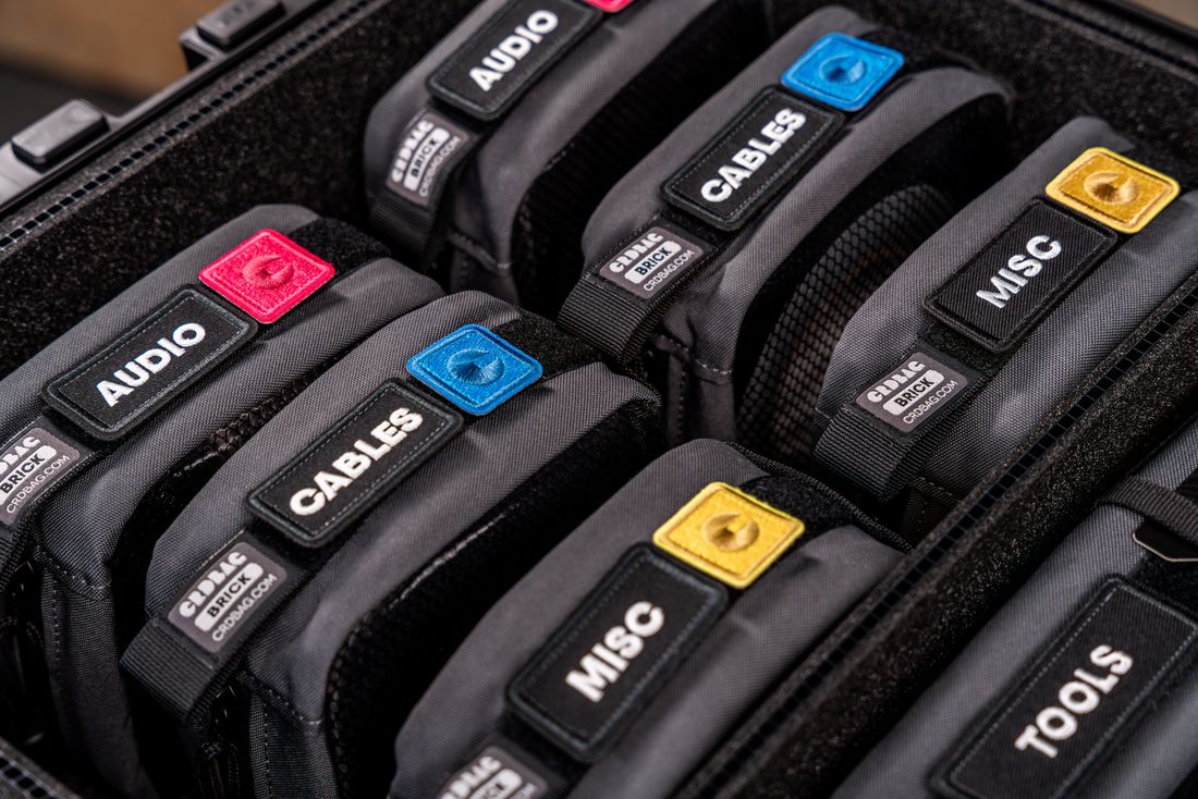 Close-up of labeled equipment pouches, marked audio, cables, misc, and tools, packed neatly inside an open hard case, nobody in frame.