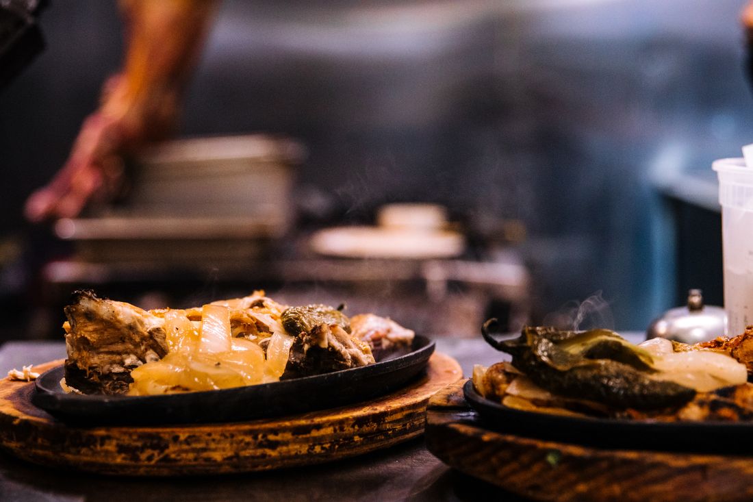 A sizzling plate of caramelized onions and roasted peppers on a cast-iron platter, steam rising, with a hand blurred at a prep station in the warm-lit kitchen behind it.