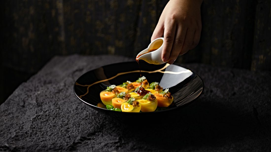 A hand pours dark sauce from a small pitcher over a plated dish on a black plate, caught mid-motion against a dim, softly lit restaurant backdrop.