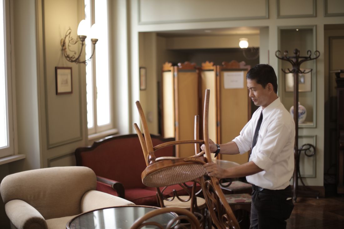 A staff member in a dress shirt and tie arranges chairs in a sunlit dining room before service, with soft window light filling the room.