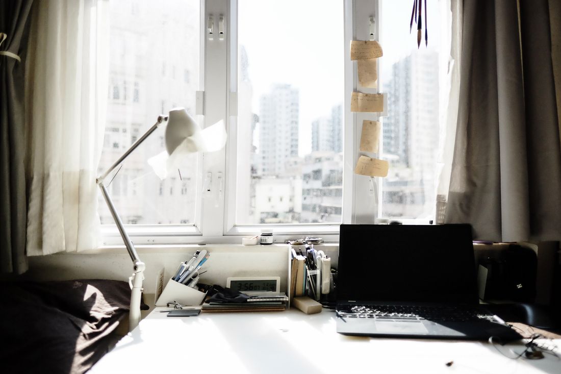 A home workspace beside a bright window, with a desk lamp, a closed laptop, pens and small paper notes clipped by the glass
