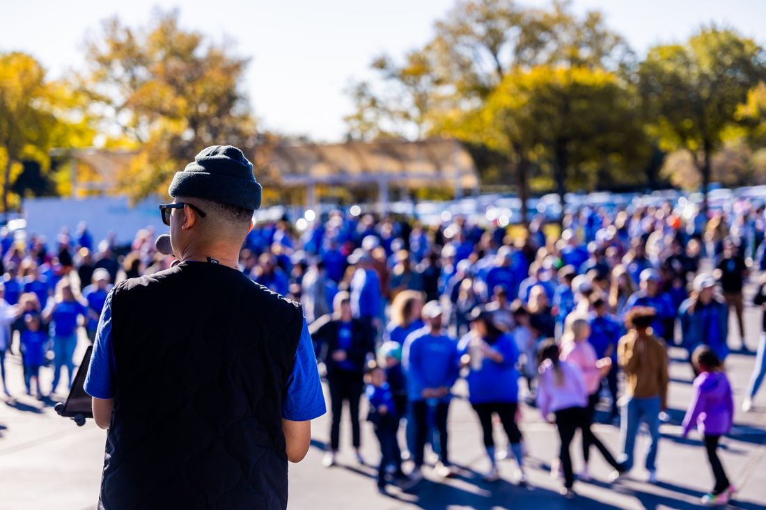 A person seen from behind speaks to a crowd standing outdoors in bright daylight, listeners facing them across an open paved area.